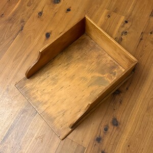 May include: A vintage wooden dustpan with a rectangular base and raised sides. The dustpan is made of light-colored wood with visible grain and a slightly worn appearance. The handle has a curved design.
