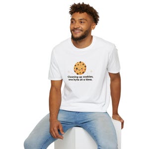 May include: White t-shirt featuring a cartoon cookie with a smiling face and chocolate chips. The text "Cleaning up cookies, one byte at a time" is printed below the cookie. The shirt is worn by a person.