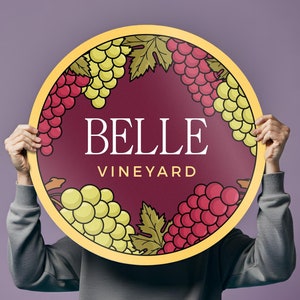 May include: A person holds a round sign with a burgundy background and a yellow border. The sign features a design of grapes and the text "BELLE VINEYARD".