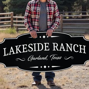 May include: A black and white sign that reads "LAKESIDE RANCH Garland, Texas" is held by a person. The sign has a decorative border. The person is wearing a plaid shirt and jeans. The background is a rural landscape.