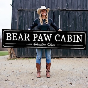 May include: A black and white sign with the text "BEAR PAW CABIN" and the location "Houston, Texas". A woman in a white cowboy hat and blue jeans is holding the sign in front of a wooden barn.