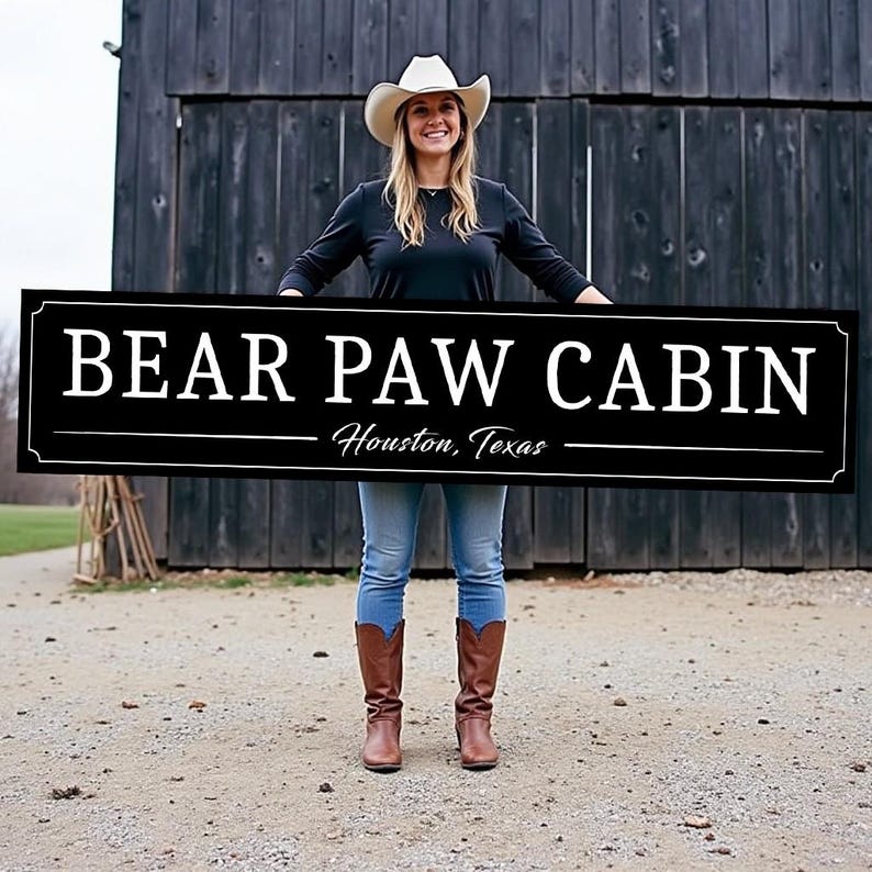 May include: A black and white sign with the text "BEAR PAW CABIN" and the location "Houston, Texas". A woman in a white cowboy hat and blue jeans is holding the sign in front of a wooden barn.