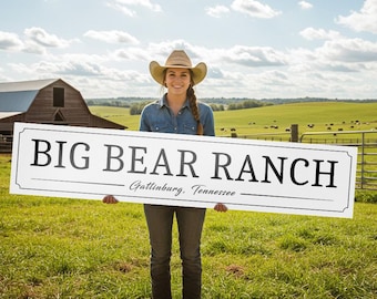 May include: A woman in a cowboy hat holds a white sign that reads "BIG BEAR RANCH, Gatlinburg, Tennessee." The sign is rectangular with a black border. The background includes a barn, green fields, and a blue sky.