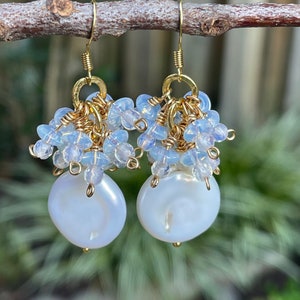 May include: Elegant gold-tone earrings featuring large, lustrous white coin pearls. Delicate clusters of light blue beads and smaller white beads adorn the top, connected by gold wire. The earrings hang from gold-colored ear wires, showcasing a sophisticated design.