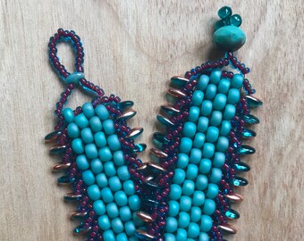 May include: A turquoise and burgundy beaded bracelet with a gold-toned accent. The bracelet has a unique, geometric design.