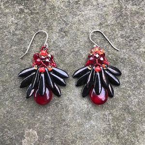 Puede incluir: Un par de pendientes colgantes con cuentas negras, rojas y naranjas. Los pendientes tienen forma de lágrima y están hechos con una variedad de cuentas, incluyendo algunas que son facetadas.
