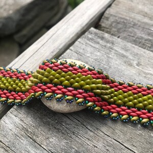 May include: A close-up of a beaded bracelet with alternating bands of red and green beads. The bracelet is lying on a wooden surface with a small rock underneath it.