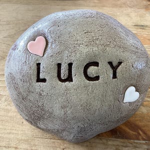 May include: A gray stone with the name "LUCY" carved into it. There are two small hearts, one pink and one white, attached to the stone.