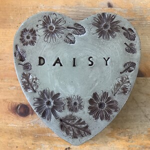 Puede incluir: Una piedra de jard&iacute;n de hormig&oacute;n en forma de coraz&oacute;n con un dise&ntilde;o en relieve de margaritas y hojas. La palabra "DAISY" est&aacute; grabada en el centro.