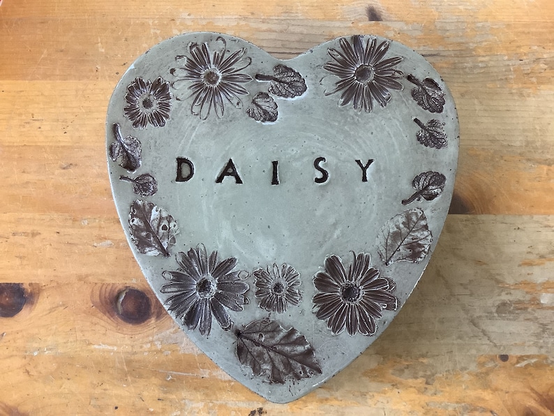 Puede incluir: Una piedra de jard&iacute;n de hormig&oacute;n en forma de coraz&oacute;n con un dise&ntilde;o en relieve de margaritas y hojas. La palabra "DAISY" est&aacute; grabada en el centro.