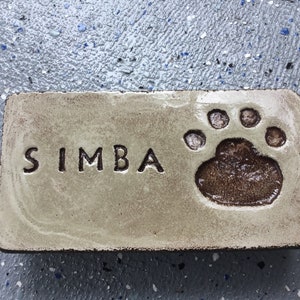 May include: A light beige ceramic tile with the name "SIMBA" engraved in black lettering and a brown paw print.