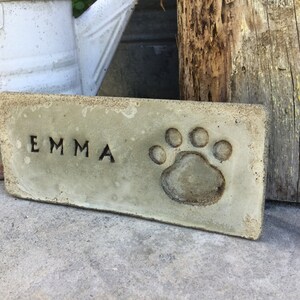 May include: A rectangular concrete plaque with the name "EMMA" engraved in black lettering and a paw print embossed in the center.