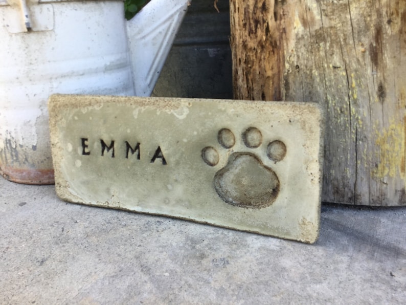 May include: A rectangular, light green concrete memorial plaque with the name "EMMA" engraved in black lettering and a paw print in the center.