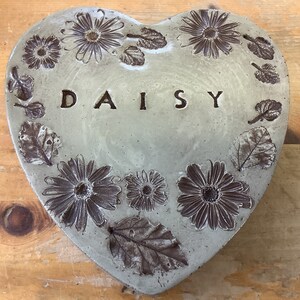 Puede incluir: Un adoqu&iacute;n de jard&iacute;n en forma de coraz&oacute;n de hormig&oacute;n con un dise&ntilde;o de margarita en relieve y la palabra "DAISY" inscrita en el centro.