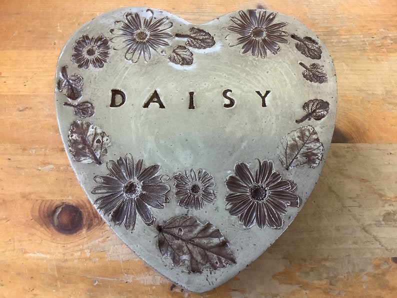 Puede incluir: Un adoqu&iacute;n de jard&iacute;n en forma de coraz&oacute;n de hormig&oacute;n con un dise&ntilde;o de margarita en relieve y la palabra "DAISY" inscrita en el centro.