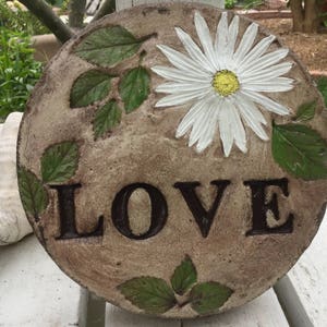 Puede incluir: Un paso de jardín redondo de piedra marrón con una margarita blanca y hojas verdes pintadas en él. La palabra "LOVE" está pintada en letras negras en la piedra.