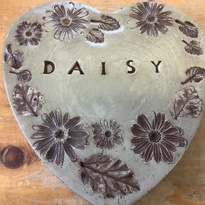 Puede incluir: Un adoqu&iacute;n de jard&iacute;n de hormig&oacute;n en forma de coraz&oacute;n con un dise&ntilde;o en relieve de margaritas y hojas. La palabra "DAISY" est&aacute; en relieve en el centro.