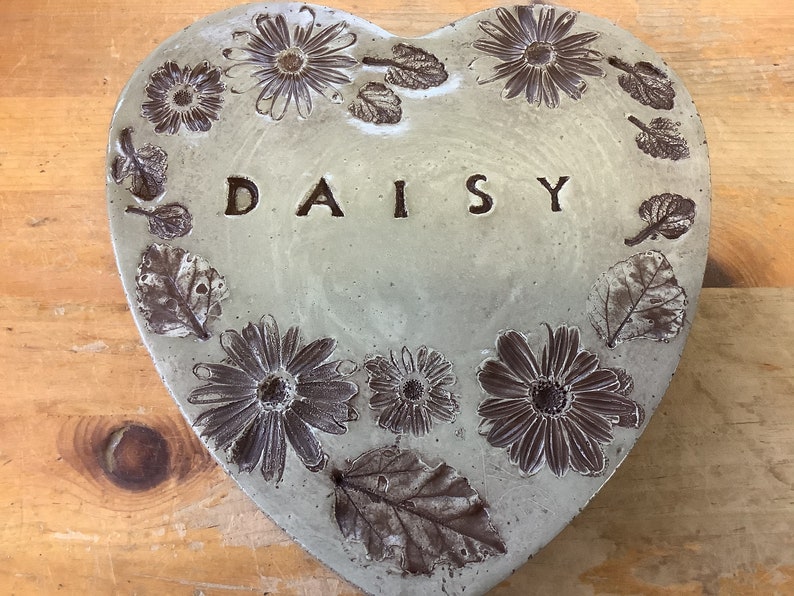 Puede incluir: Un adoqu&iacute;n de jard&iacute;n de hormig&oacute;n en forma de coraz&oacute;n con un dise&ntilde;o en relieve de margaritas y hojas. La palabra "DAISY" est&aacute; en relieve en el centro.