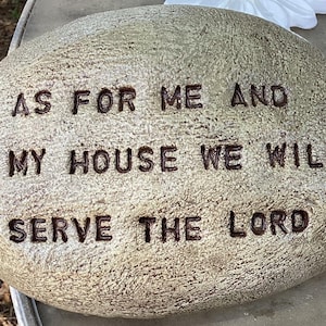 May include: A decorative, oval-shaped stone with the inscription "AS FOR ME AND MY HOUSE WE WILL SERVE THE LORD" carved into its surface. The stone has a textured, light brown appearance.