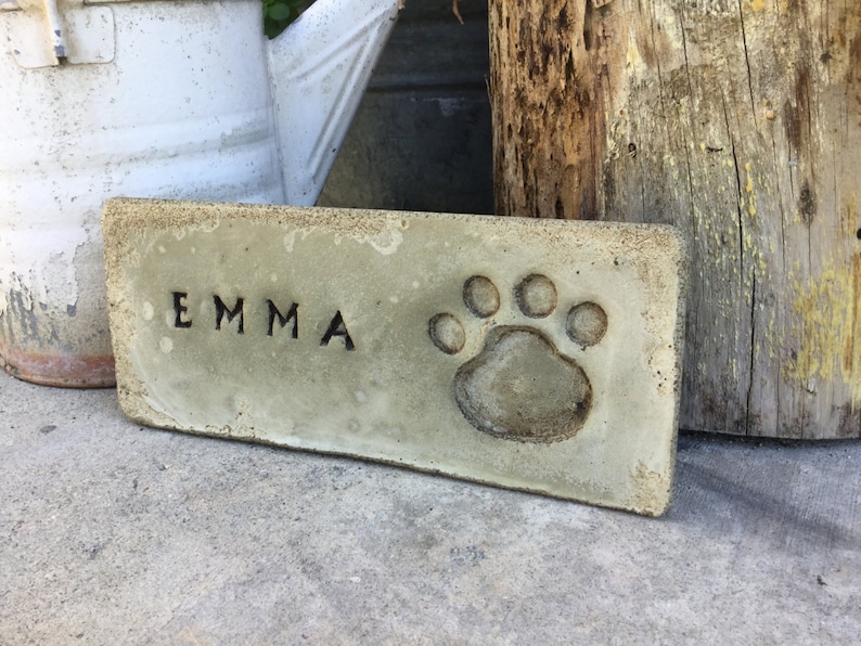 May include: A rectangular concrete plaque with the name "EMMA" engraved in black lettering and a paw print embossed in the center.
