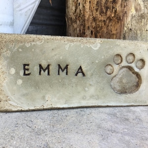 May include: A light gray concrete rectangular plaque with the name "EMMA" engraved in black lettering and a paw print in the upper right corner.