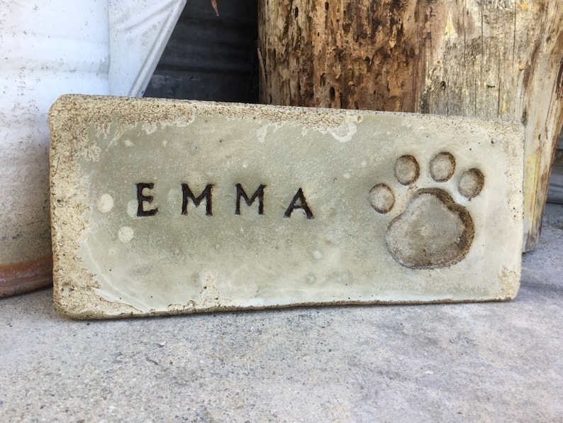 May include: A light gray concrete rectangular plaque with the name "EMMA" engraved in black lettering and a paw print in the upper right corner.