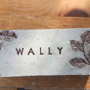 May include: A white rectangular concrete plaque with the word "WALLY" embossed in the center. The plaque has two leaf impressions on the left side and a branch with three leaf impressions on the right side.