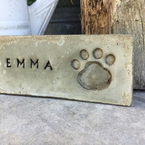 May include: A light green concrete memorial brick with the name "EMMA" and a paw print.