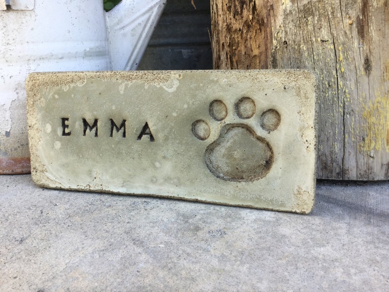 May include: A light green concrete memorial brick with the name "EMMA" and a paw print.