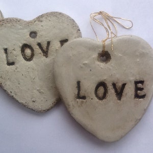 Puede incluir: Dos adornos de corazón de cerámica blanca con la palabra "LOVE" escrita en ellos. Un corazón tiene una cuerda dorada atada a él.