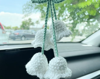May include: A white crocheted flower car charm with green leaves hanging from the rearview mirror of a car. The charm is made of yarn and has a delicate, feminine design.