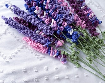May include: A bunch of crocheted lavender flowers in shades of purple, pink, and white. The flowers are arranged in a bouquet and have green stems.