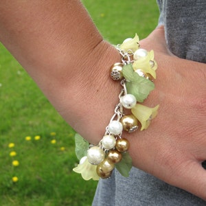 Peut inclure: Un bracelet à breloques argenté avec des feuilles vertes, des perles blanches et des breloques de fleurs jaunes.