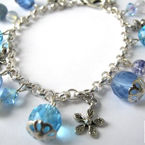 May include: Silver-toned charm bracelet featuring light blue faceted beads, round blue beads, and snowflake charms. The bracelet includes various sizes and shapes of blue and clear beads, creating a delicate and elegant accessory.