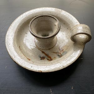 May include: A speckled ceramic candle holder with a circular base and a central cup for the candle. It has a small handle on the side. The color is a mix of beige and brown, with darker brown accents.