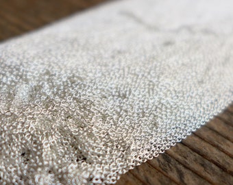May include: Close-up of a length of delicate silver chain, composed of numerous small, interconnected links. The chain is displayed against a blurred wooden surface, creating a contrast between the shiny metal and the warm wood tones. The chain appears to be new.