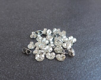 May include: A collection of shiny silver earring backs scattered on a dark gray surface. The earring backs are small, with a butterfly-shaped design and a post for securing earrings. The backs are reflective, catching the light.