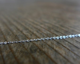 May include: A delicate silver chain necklace with small, evenly spaced links. The chain is displayed against a blurred, textured wooden surface, creating a contrast between the shiny metal and the rustic background. The focus is on the chain's intricate design.