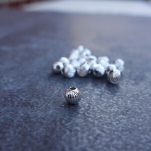 May include: A collection of shiny, silver-colored beads with a ribbed design. The beads are round with a small hole at the top. The beads are scattered on a dark gray surface.