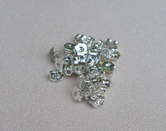 May include: A collection of shiny silver earring backs, scattered on a neutral-toned surface. The earring backs have a flower-like design and are made of a reflective metal, likely sterling silver.