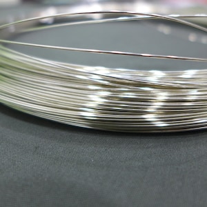 Puede incluir: Primer plano de una bobina de alambre brillante de color plateado. El alambre está enrollado firmemente, creando una apariencia densa y texturizada. El alambre parece ser de metal y probablemente se utiliza para manualidades o joyería.