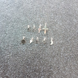 May include: A selection of small, silver-coloured charms in diverse shapes, including a circle, star, cross, square, heart, and crescent moon. Each charm has a small loop for attaching to jewellery. The charms are displayed on a grey surface.