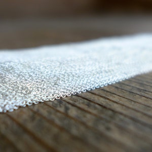 May include: Close-up of a collection of delicate silver chain links. The chains are arranged in a row, with a blurred wooden surface in the background. The chains are small and shiny, reflecting light.