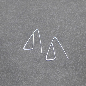 Puede incluir: Dos pendientes minimalistas de metal blanco. Cada pendiente presenta un diseño triangular con una línea recta que se extiende desde la parte superior. Los pendientes se muestran sobre una superficie gris.