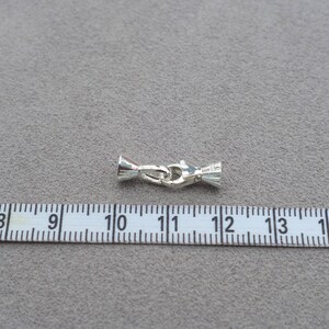Può includere: Chiusura per gioielli color argento, lunga circa 2 cm, con un design decorativo. La chiusura è posizionata su una superficie grigia accanto a un righello. La chiusura ha una forma a barilotto con chiusura a gancio.