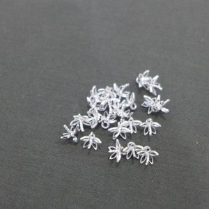 May include: A collection of small, silver-colored flower-shaped charms. These charms have a delicate, openwork design with five petals and a small loop for attaching to jewelry or other crafts. The charms are scattered on a dark gray surface.