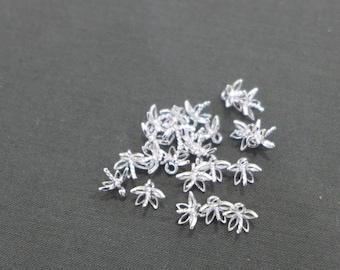 May include: A collection of small, silver-colored flower-shaped charms. These charms have a delicate, openwork design with five petals and a small loop for attaching to jewelry or other crafts. The charms are scattered on a dark gray surface.