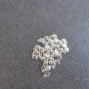 May include: A collection of small, silver-coloured heart charms with a loop at the top for stringing. The charms are scattered on a dark grey surface. These charms are ideal for jewellery making or craft projects.