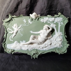 May include: A green and white porcelain wall plaque with a raised relief design of a woman reclining in a forest setting with a cherub flying above her. The plaque has a decorative, ornate frame.