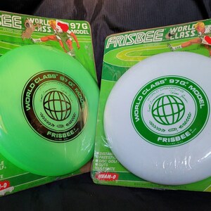 May include: Two unopened packages of Wham-O World Class 97g model frisbees. One frisbee is green and the other is white. Both frisbees have a black and green logo with the text "World Class 97g Model" and "Frisbee" printed on them.